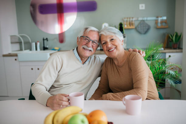 Portrait of a happy married senior couple enjoying themselves at home with a cup of coffee, sitting at the dining table in their kitchen and looking at the camera